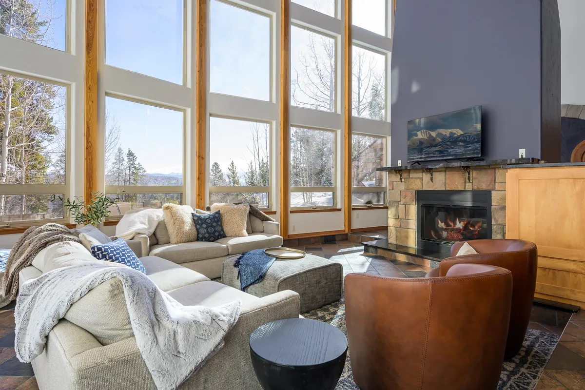 A warm and welcoming living area with comfortable seating arranged by a glowing fireplace, large windows with scenic views, and decorative pillows that add a touch of style. Sunlight streams in, enhancing the sense of space and togetherness.