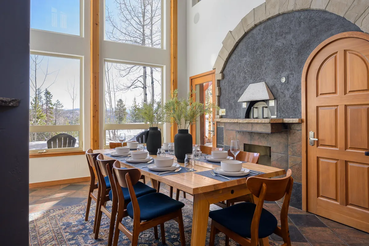 A dining area adjacent to large windows, featuring a substantial dining table set for a group, a modern fireplace, and vases with greenery. The open layout and visual connection to the outdoors create a space ideal for shared meals and celebrations.