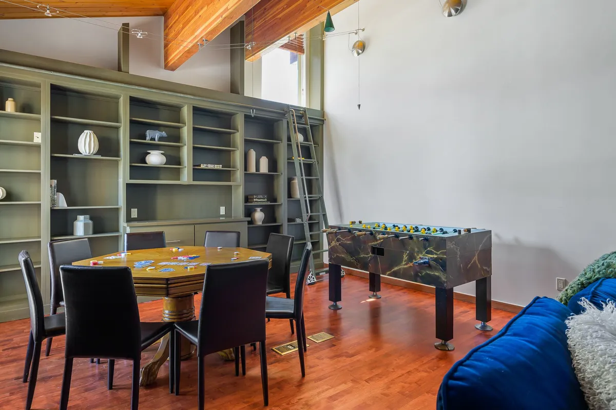 This image features a vibrant and playful game room with a foosball table, a dining table set up with board games, shelves decorated with books and accessories, and large windows that let in plenty of natural light. The room feels lively and welcoming, perfectly suited for group games and family gatherings.