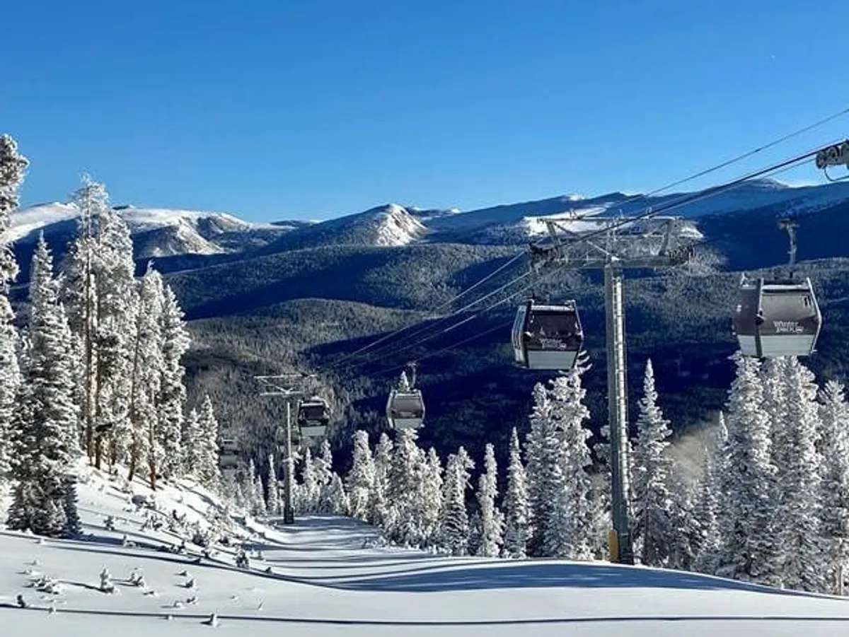 A serene winter landscape featuring a ski lift ascending through snow-covered trees, with a dramatic mountain range in the background. The scene highlights fresh powder snow and the anticipation of alpine adventures.