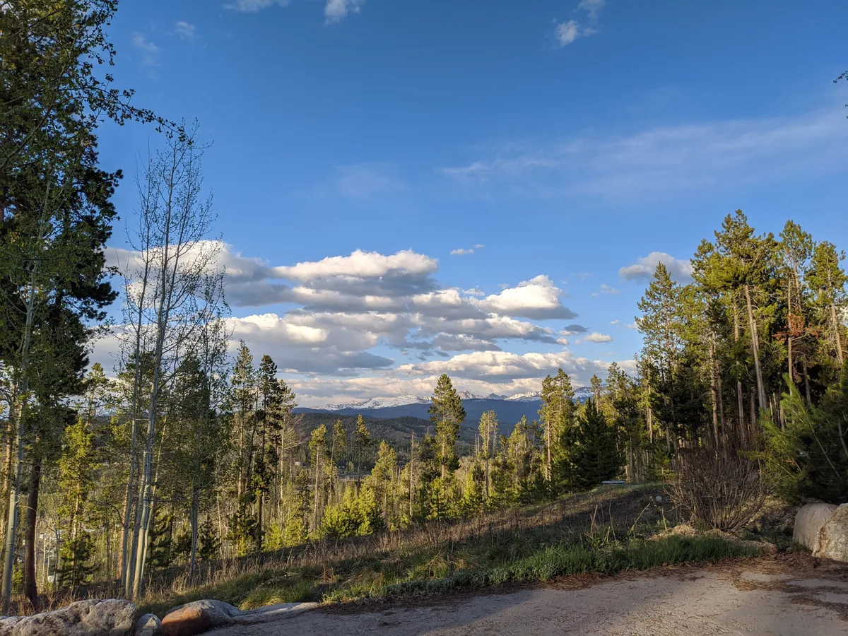 A wide, scenic view of distant mountains, tall trees, and lush green landscapes under an expansive sky. The tranquility and openness of the setting are ideal for outdoor activities and relaxation, with a clear sense of Colorado’s natural beauty.