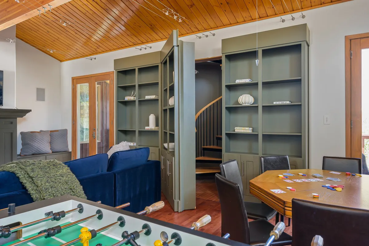 A multi-use living space featuring a blue sofa, dining table, foosball table, bookshelves, and a striking spiral staircase leading to a loft. The eclectic design invites activities like board games, socializing, reading, and movie watching—all key aspects mentioned in the text.