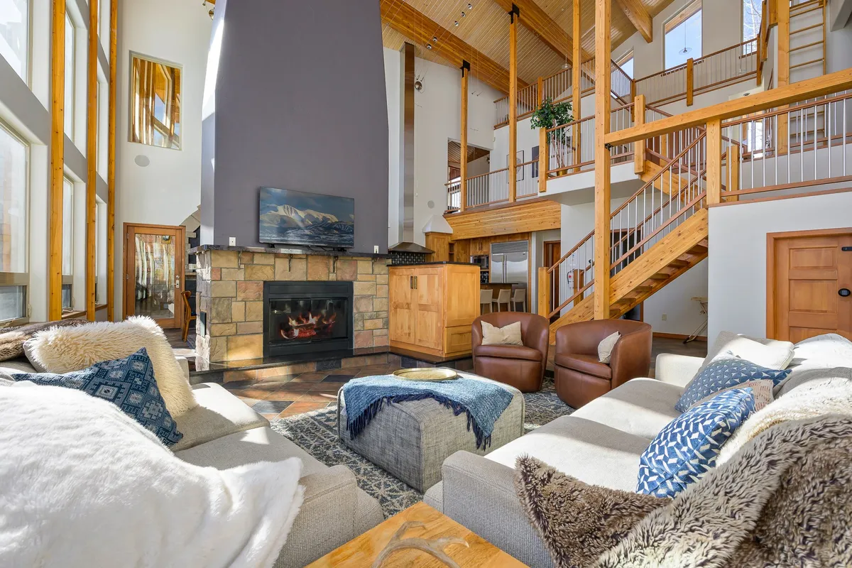 A spacious great room featuring a prominent gas fireplace, large windows that let in abundant natural light, plush sofas arranged for conversation, and an inviting staircase. The windows showcase a stunning backdrop of mountains and greenery, while the seating creates a cozy, communal atmosphere.