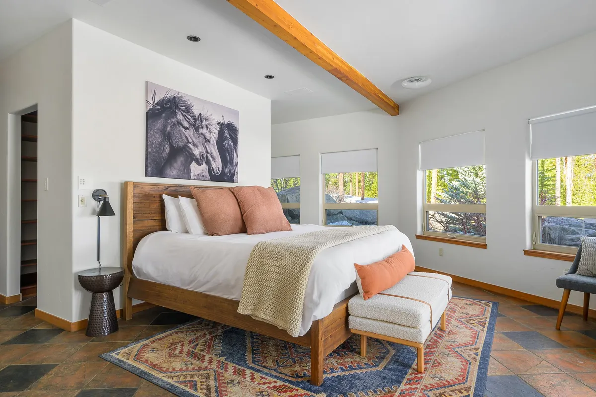 A serene bedroom featuring a plush bed with soft pillows, horses artwork above the headboard, rustic wooden beams, and an area rug. Sunlight streams in through nearby windows, casting a calm glow over the cozy space.