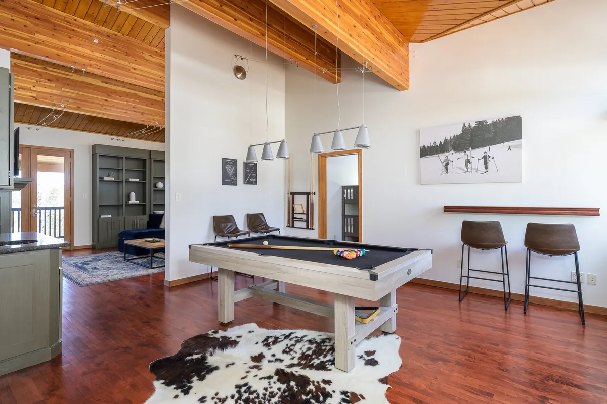 A spacious lounge centered on a classic pool table, with wooden beams overhead, bar stools nearby for spectators, and colorful artwork on the wall. The space is designed for friendly competitions and casual socializing.